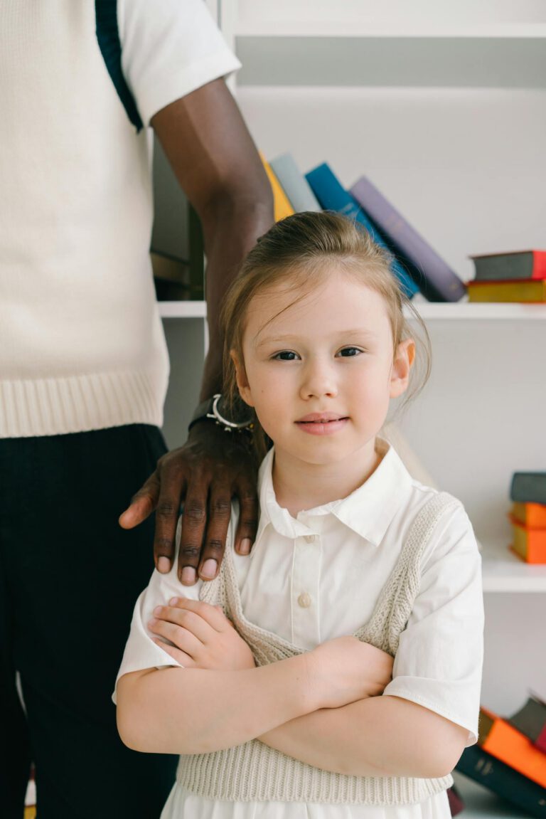 Charming young girl standing confidently with books on a shelf behind.